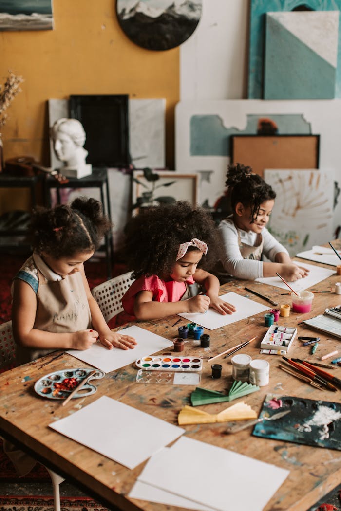 About Three young girls engaged in painting at an indoor art workshop, using watercolors and brushes.